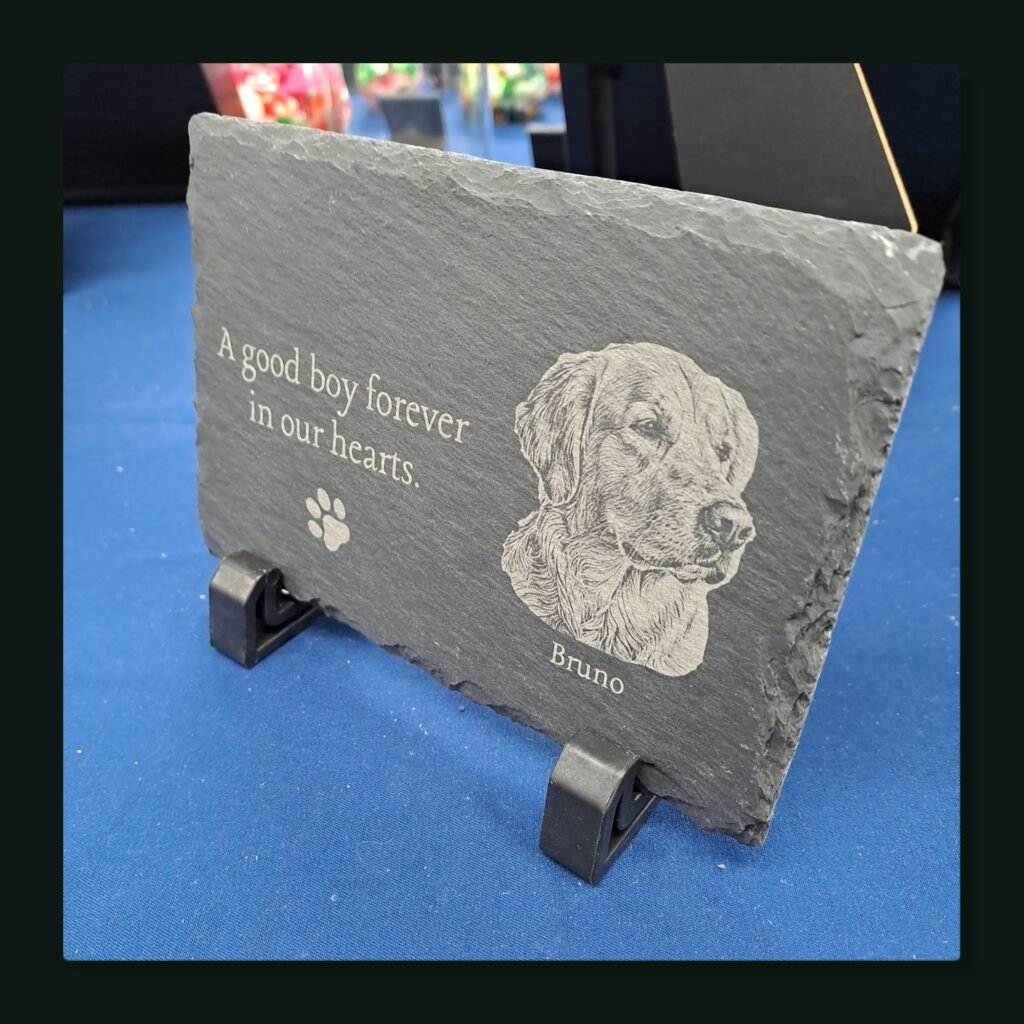Personalised slate dog memorial plaque engraved with a Golden Retriever portrait, paw print, text "A good boy forever in our hearts", named Bruno, resting on black 3D printed display stands.