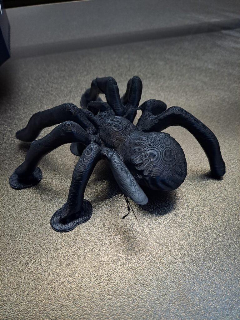 A 3D-printed black spider model resting on a textured surface, showing visible layer lines, detailed legs, and a missing front leg.