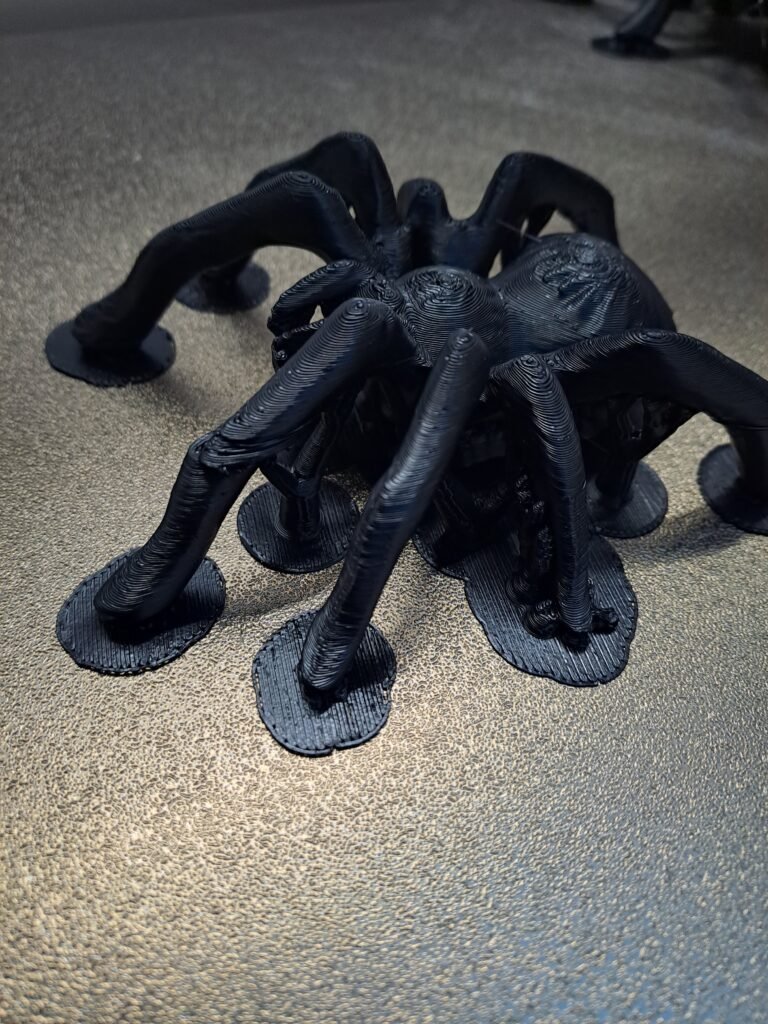 Black 3D printed spider model sitting on a textured print bed with a failed print leg on the right side.